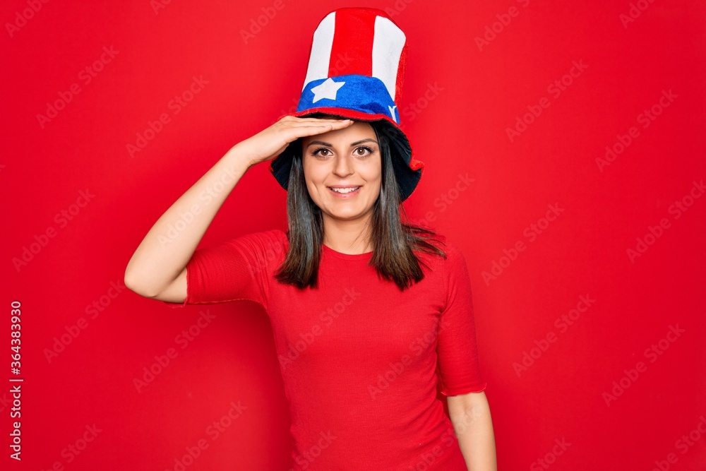 Young beautiful brunette woman wearing united states hat celebrating independence day very happy and smiling looking far away with hand over head. Searching concept. Young beautiful brunette woman wearing united states hat celebrating independence day very happy and smiling looking far away with hand over head. Searching concept.