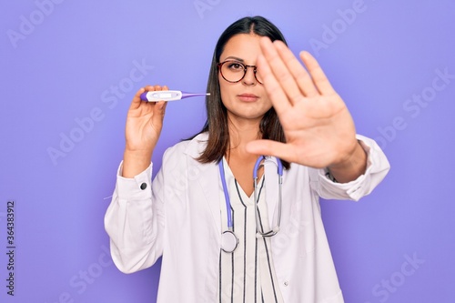 Young beautiful brunette doctor woman wearing stethoscope and coat holding thermometer with open hand doing stop sign with serious and confident expression, defense gesture