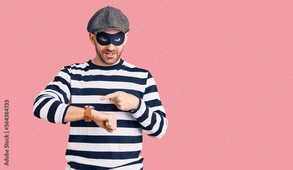 Young handsome man wearing burglar mask in hurry pointing to watch time, impatience, upset and angry for deadline delay Young handsome man wearing burglar mask in hurry pointing to watch time, impatience, upset and angry for deadline delay