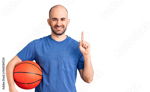 Young handsome man holding basketball ball surprised with an idea or question pointing finger with happy face, number one © Krakenimages.com