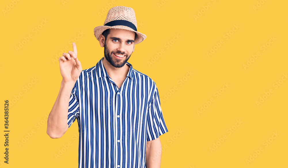 Young handsome man with beard wearing summer hat and striped shirt showing and pointing up with finger number one while smiling confident and happy.