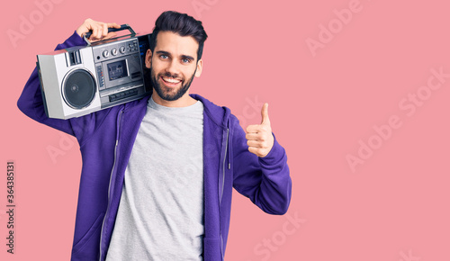 Young handsome man with beard listening to music using vintage boombox smiling happy and positive, thumb up doing excellent and approval sign