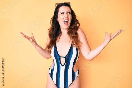 Young beautiful brunette woman wearing summer swimwear and sunglasses crazy and mad shouting and yelling with aggressive expression and arms raised. frustration concept.