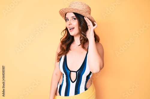 Young beautiful brunette woman wearing bikini and summer hat smiling with hand over ear listening and hearing to rumor or gossip. deafness concept.