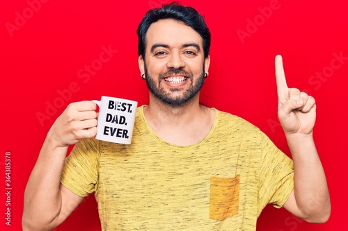 Young hispanic man drinking mug of coffe with best dad ever message smiling with an idea or question pointing finger with happy face, number one photo