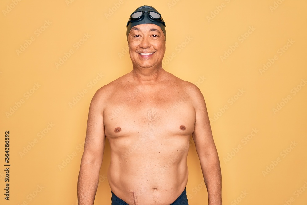 Middle age senior grey-haired swimmer man wearing swimsuit, cap and goggles with a happy and cool smile on face. Lucky person.