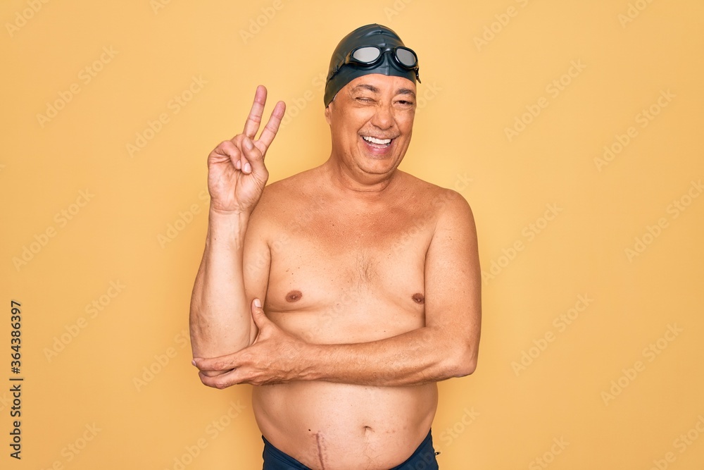 Middle age senior grey-haired swimmer man wearing swimsuit, cap and goggles smiling with happy face winking at the camera doing victory sign with fingers. Number two.