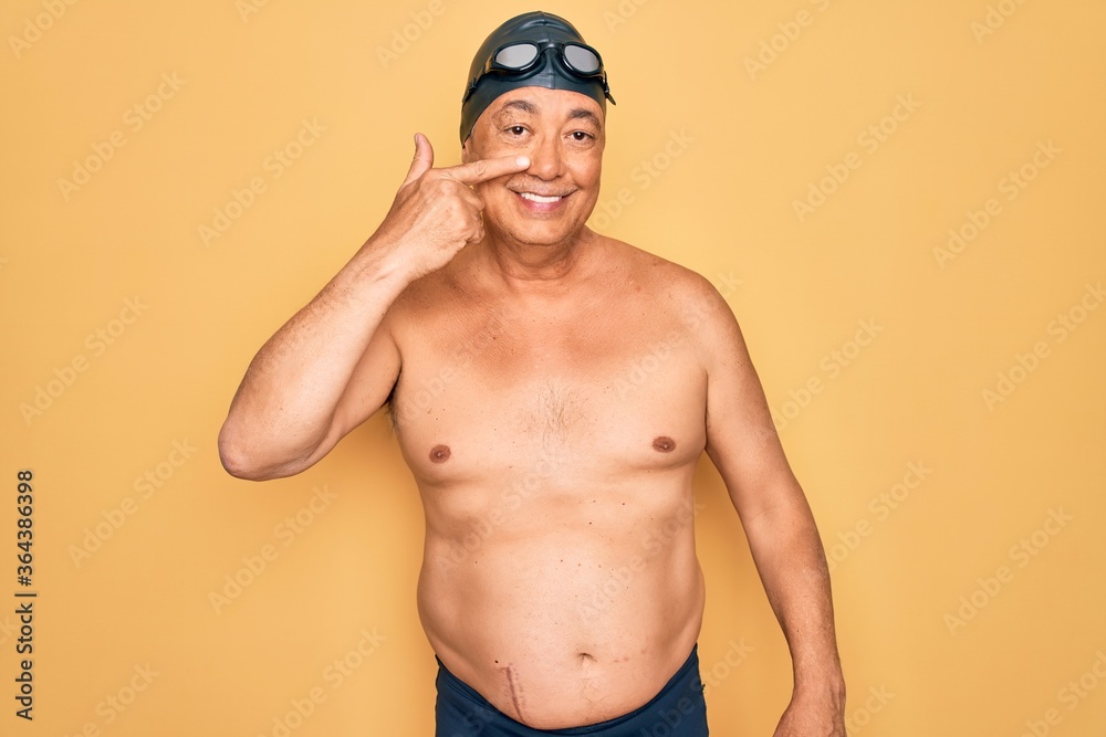 Middle age senior grey-haired swimmer man wearing swimsuit, cap and goggles Pointing with hand finger to face and nose, smiling cheerful. Beauty concept