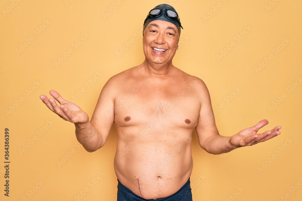 Middle age senior grey-haired swimmer man wearing swimsuit, cap and goggles smiling cheerful with open arms as friendly welcome, positive and confident greetings