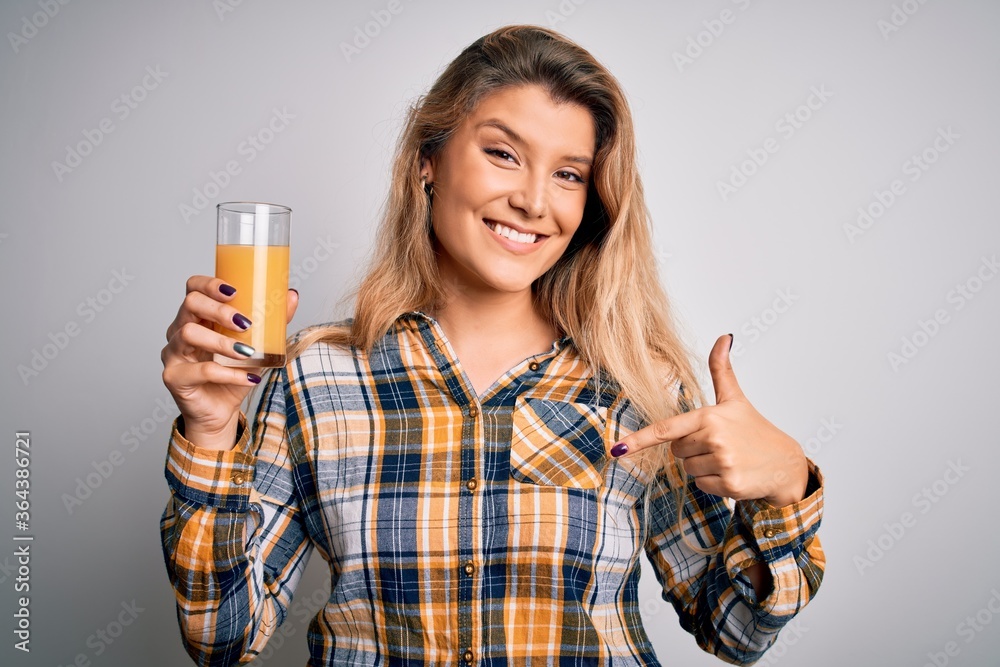 Young beautiful blonde woman drinking glass of healthy orange juice over white background with surprise face pointing finger to himself Young beautiful blonde woman drinking glass of healthy orange juice over white background with surprise face pointing finger to himself