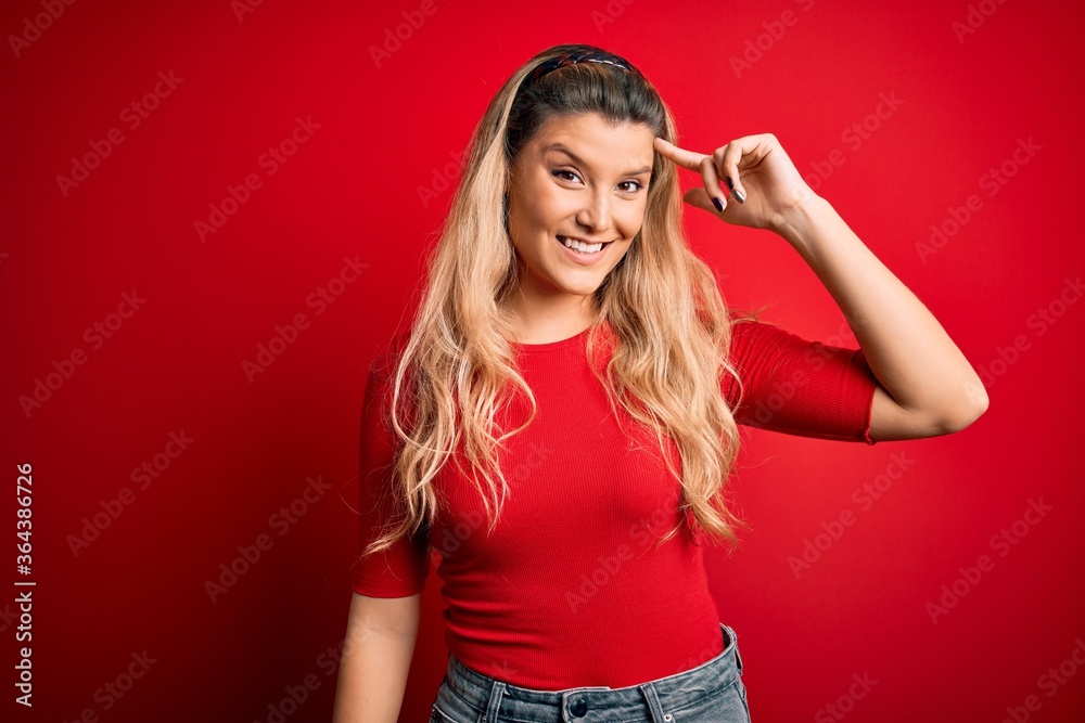 Young beautiful blonde woman wearing casual t-shirt standing over isolated red background Smiling pointing to head with one finger, great idea or thought, good memory