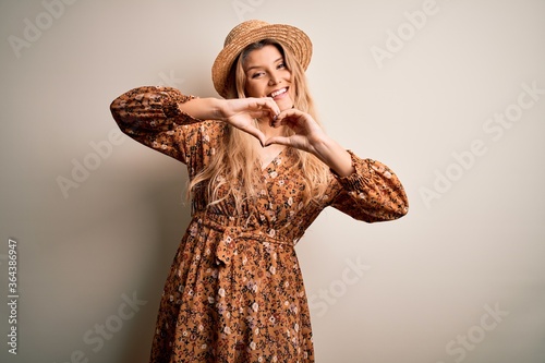 Young beautiful blonde woman wearing summer dress and hat over isolated white background smiling in love doing heart symbol shape with hands. Romantic concept.