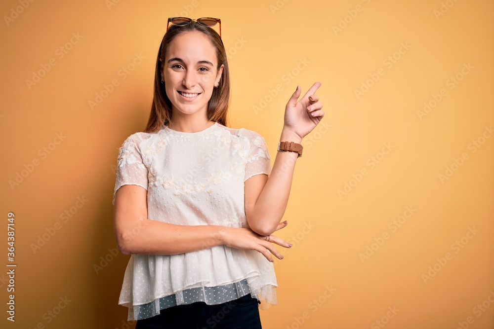 Young beautiful woman wearing casual t-shirt and sunglasses over isolated yellow background with a big smile on face, pointing with hand and finger to the side looking at the camera. Young beautiful woman wearing casual t-shirt and sunglasses over isolated yellow background with a big smile on face, pointing with hand and finger to the side looking at the camera.