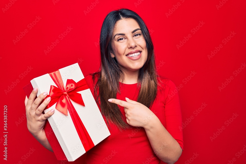 Young beautiful brunette woman holding birthday gift over isolated red background smiling happy pointing with hand and finger Young beautiful brunette woman holding birthday gift over isolated red background smiling happy pointing with hand and finger