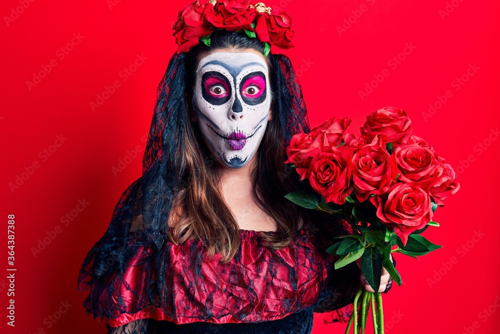 Young woman wearing day of the dead costume holding flowers scared and amazed with open mouth for surprise, disbelief face Young woman wearing day of the dead costume holding flowers scared and amazed with open mouth for surprise, disbelief face