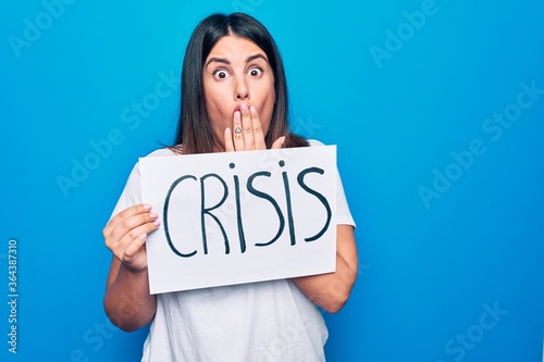 Young beautiful brunette woman holding banner with crisis message over blue background covering mouth with hand, shocked and afraid for mistake. Surprised expression