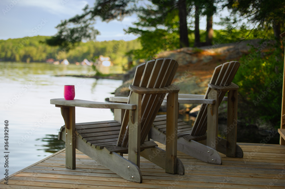 Two Adirondack chairs sitting on a cottage wooden dock facing the calm water of a lake in Muskoka, Ontario Canada. A purple insulated tumbler glass is visible on of of the chairs. - obrazy, fototapety, plakaty 