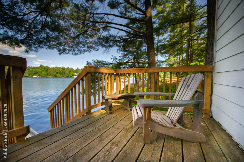 Adirondack chair sitting on a cottage wooden deck facing a calm lake during a summer day in Muskoka, Ontario Canada. - obrazy, fototapety, plakaty 