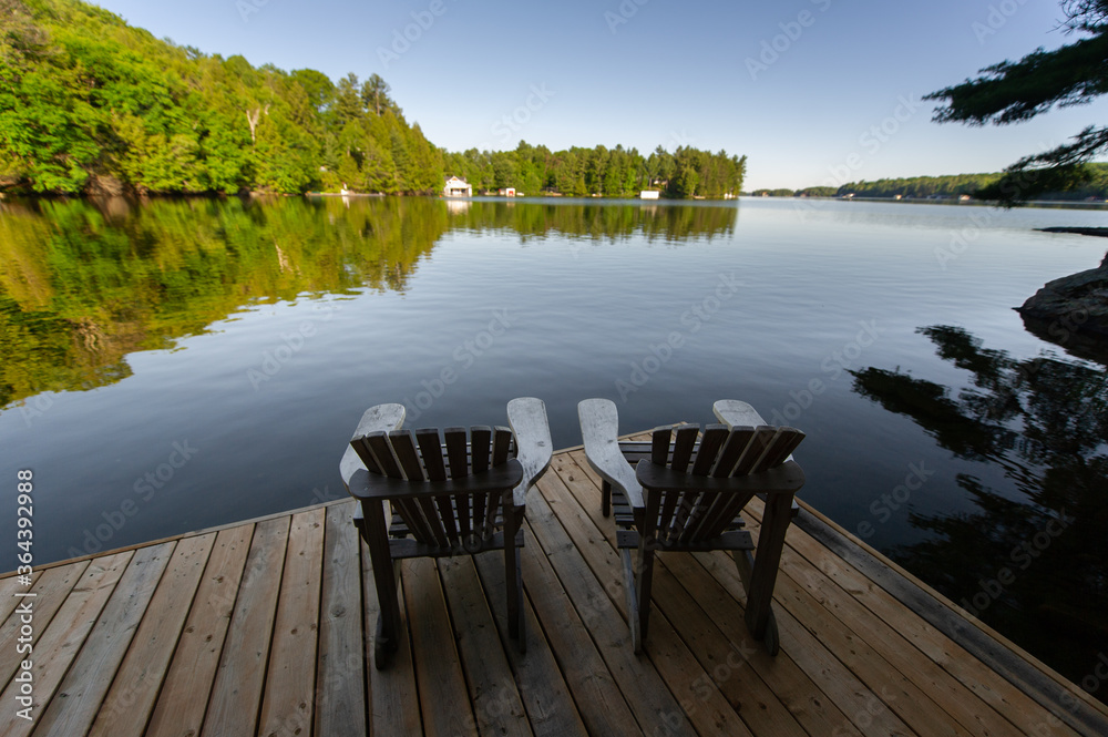 Two Adirondack chairs sitting on a wooden dock facing the calm water of a lake in Muskoka, Ontario Canada. Cottages are visible nestled between trees in background. 