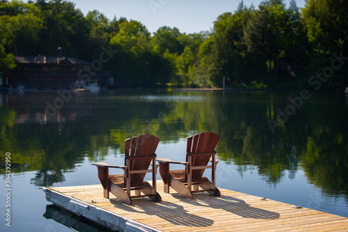 Two Adirondack chairs sitting on a wooden dock facing the calm water of a lake in Muskoka, Ontario Canada. Cottages are visible nestled between trees in background.  photo