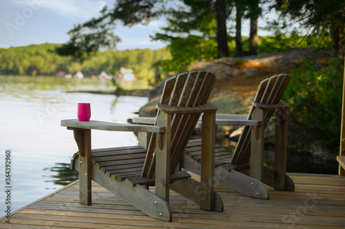 Fototapeta Two Adirondack chairs sitting on a cottage wooden dock facing the calm water of a lake in Muskoka, Ontario Canada