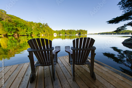 Two Adirondack chairs sitting on a wooden dock facing the calm water of a lake in Muskoka, Ontario Canada. Cottages are visible nestled between trees in background. 