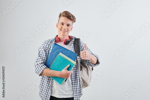 Portrait of happy handsome young high school student with textbooks showing thumbs-up and smiling at camera photo
