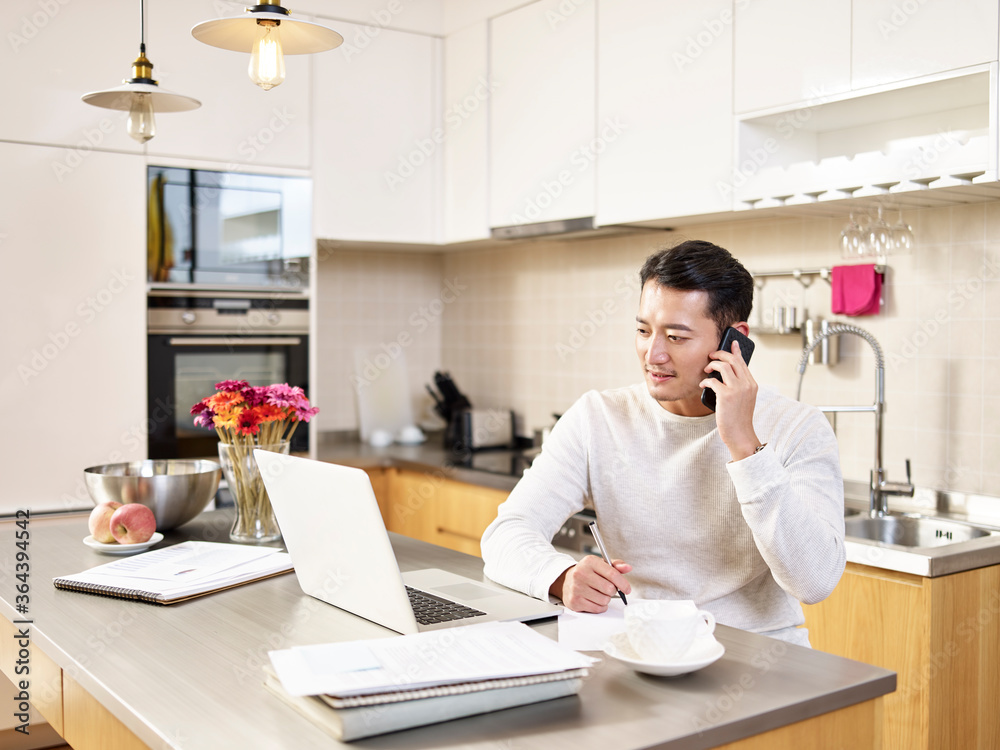 young asian business man working from home looking at laptop using talking on mobile phone