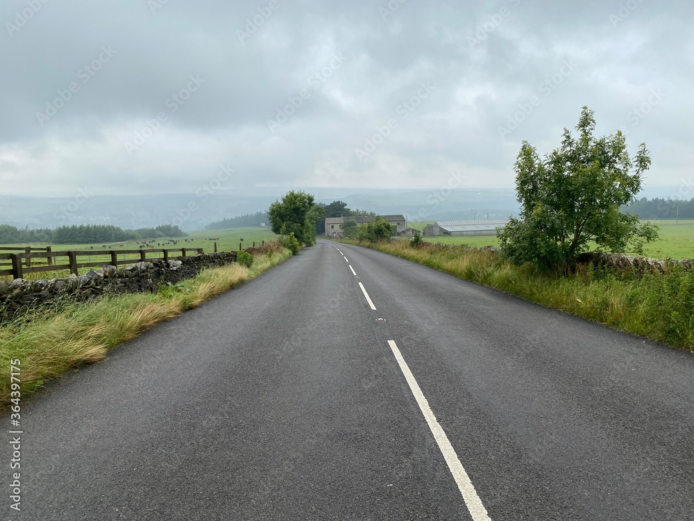 Country road with dry stone walls, trees, and fields, with heavy rain in the distance near, Slaithwaite, Oldham, UK