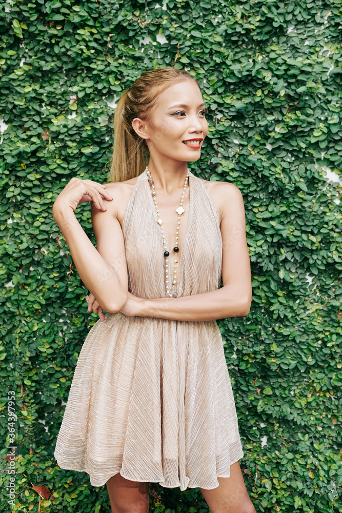 Stylish slim young woman standing at green hedge of fower garden, smiling at looking away Stylish slim young woman standing at green hedge of fower garden, smiling at looking away
