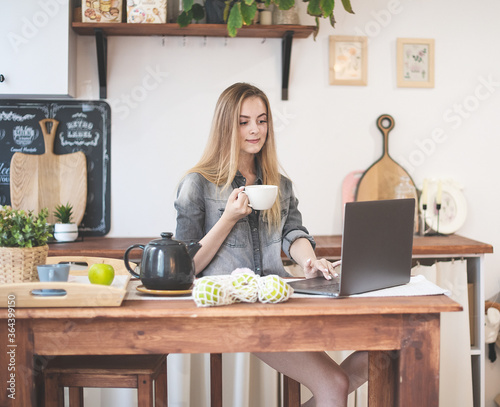 A young attractive girl with long hair has Breakfast, works and communicates on a laptop remotely in the kitchen at home. 