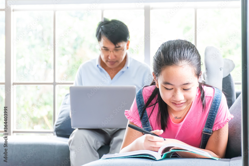 daughter reading a book and lying on sofa in living room while father work from home with laptop. asian family spending time together. daughter reading a book and lying on sofa in living room while father work from home with laptop. asian family spending time together.