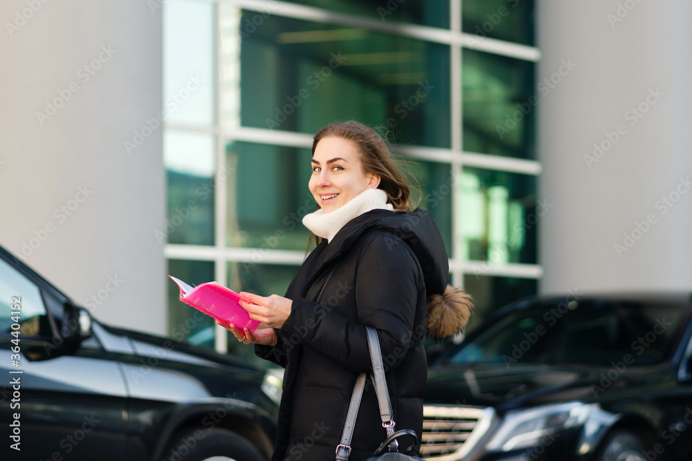 Attractive business lady in the parking lot of a business center. Positive human emotions. The girl holds a notebook, has fun and enjoys life. Good deal in the business center. Soft focus Attractive business lady in the parking lot of a business center. Positive human emotions. The girl holds a notebook, has fun and enjoys life. Good deal in the business center. Soft focus