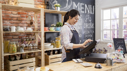 smiling coffee shop assistant using pos point of sale terminal to put in order from note paper at restaurant register. waitress lady working in counter in cafe store. young girl staff using cashbox. photo