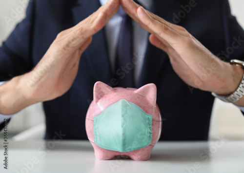 Male bank manager with piggy bank in medical protective mask sitting at table  closeup