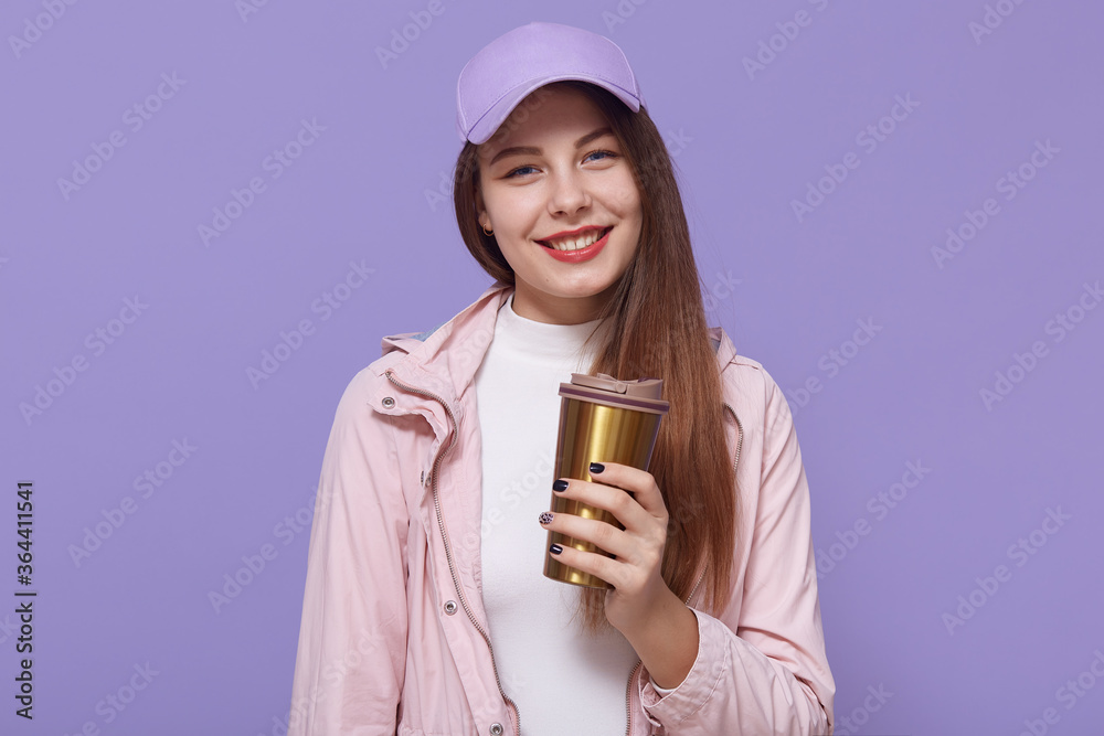 Shot of glad female student has coffee break after lectures, enjoys hot beverage from thermo mug, posing isolated over lilac background, wearing casual attire. Shot of glad female student has coffee break after lectures, enjoys hot beverage from thermo mug, posing isolated over lilac background, wearing casual attire.