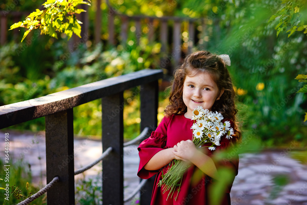 Fototapeta premium Summer portrait of a charming little girl with a bouquet of daisies in nature. A girl in a red dress holds a bouquet, smiles and looks at the camera.