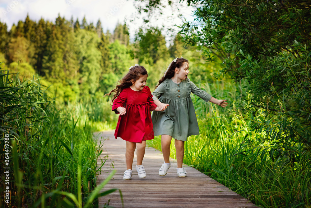 Naklejka premium Two little girls with long hair in a green and red dress run along a wooden path in the Park. A fun childhood of the two sisters.