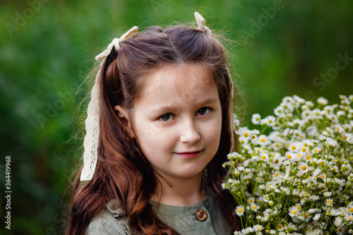 Summer portrait of a charming little girl with a bouquet of daisies in nature. A girl in a green dress holds a bouquet, smiles and looks at the camera. © Elena 
