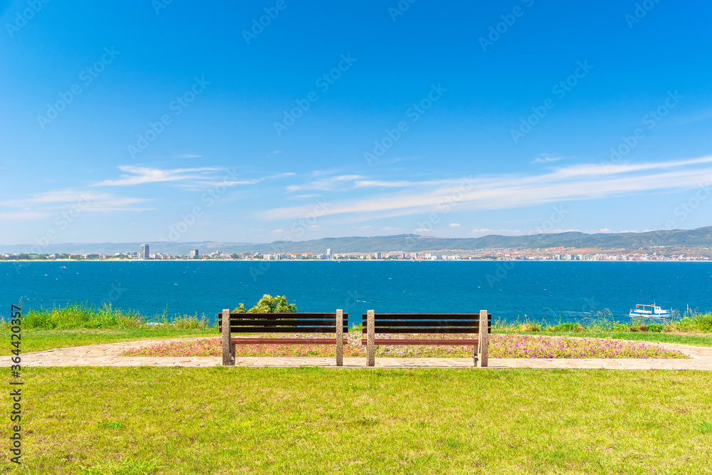two benches on the sunny beach shore. beautiful view from paved footpath on the seaside. city and mountain in the distance beneath a blue sky with clouds