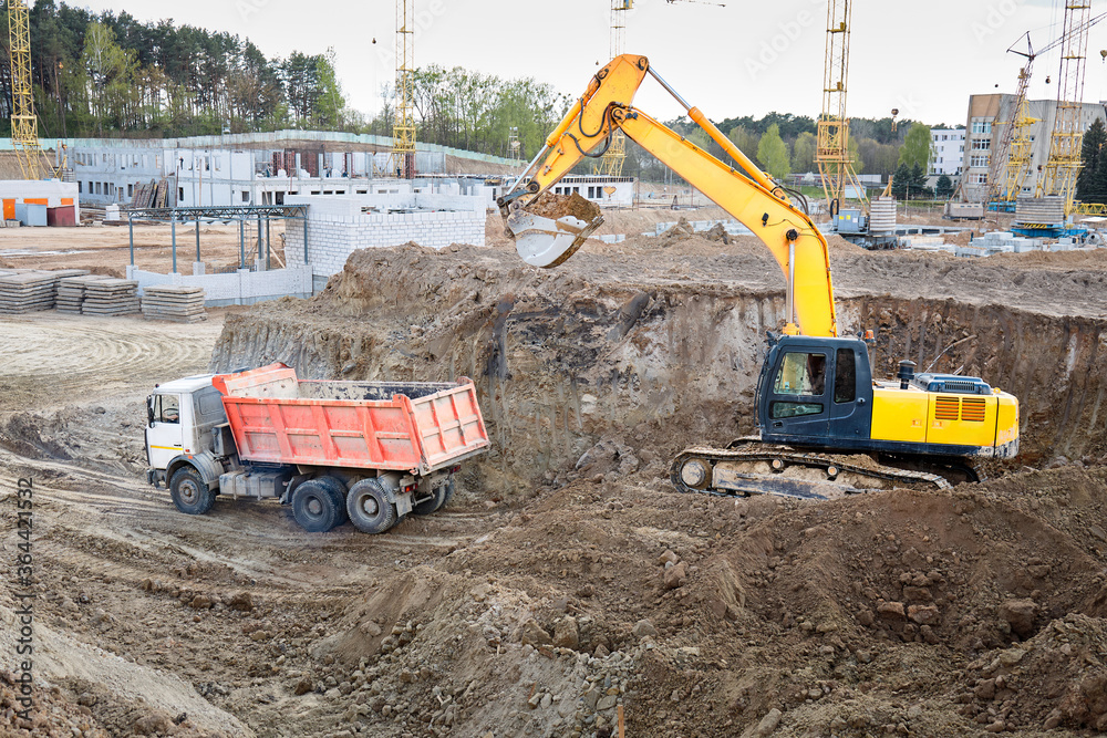 Crawler excavator in process of loading sand soil to multi-ton mining truck at construction site with cranes buildings and workers in background. Crawler excavator in process of loading sand soil to multi-ton mining truck at construction site with cranes buildings and workers in background.
