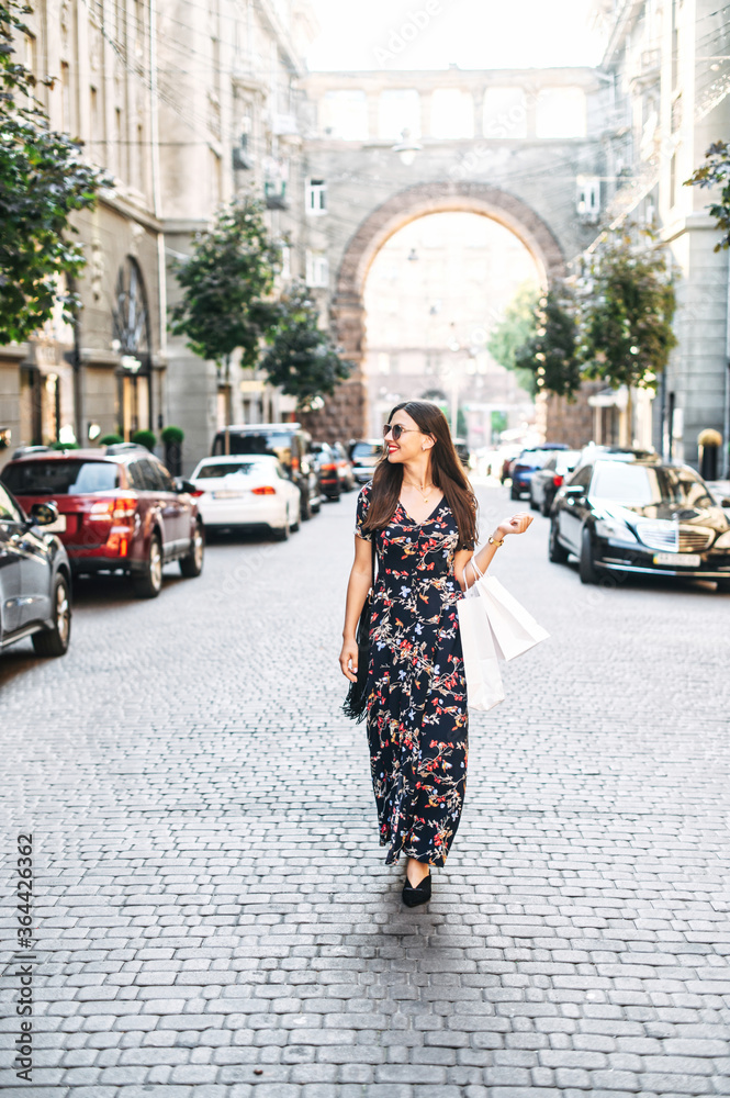 Full length young attractive woman in a long summer dress is walking down the street with a paper shopping bags in hands