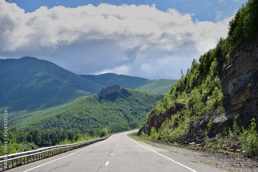 Highway among the hills. White clouds cover the green mountains. Bright summer day.