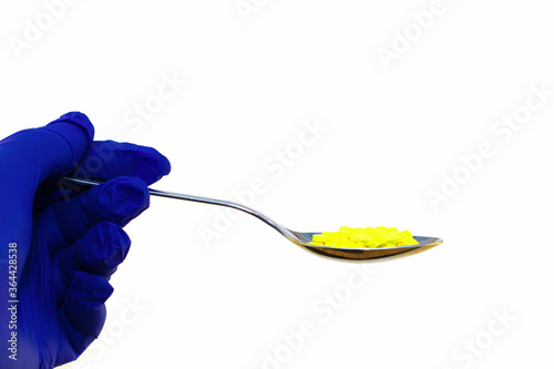 hand in a blue protective surgical glove holds an iron spoon with yellow pills on the white background