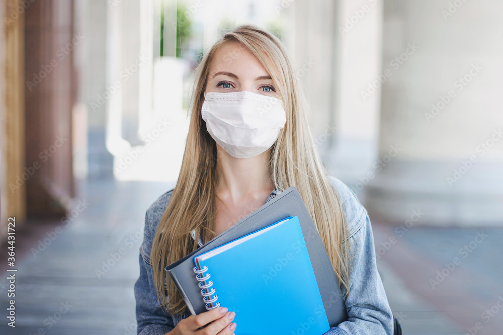 Caucasian student girl wearing a medical mask holds exercise book in her hands and looks to the camera, the new rules for education during the coronovirus quarantine, a pandemic in schools Caucasian student girl wearing a medical mask holds exercise book in her hands and looks to the camera, the new rules for education during the coronovirus quarantine, a pandemic in schools