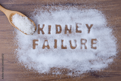 White granulated natural sea salt in wooden scoop and words" kidney failure" letters written in salt grains on wood table background. Unhealthy food concept. Top view. Flat lay.