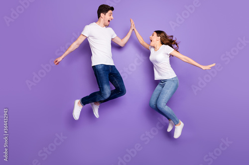 Full length profile side photo of cheerful two people freelancer jump win discount clap high five gesture wear white t-shirt denim jeans gumshoes isolated over violet color background