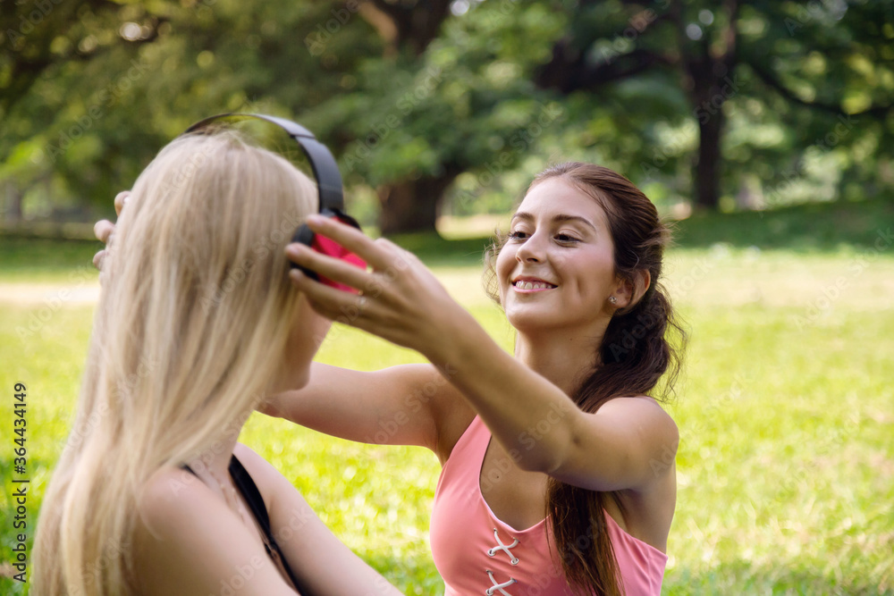 Naklejka premium smiling young beautiful teenage girl gives earphones and headphones to listen to the music to lovely blond hair friend girl in the park at the weekend. people and lifestyle concept
