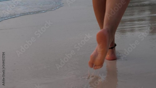 Backview of female legs walk along the sandy beach. Waves washing away foot steps at sunset. Girl with bare feet.