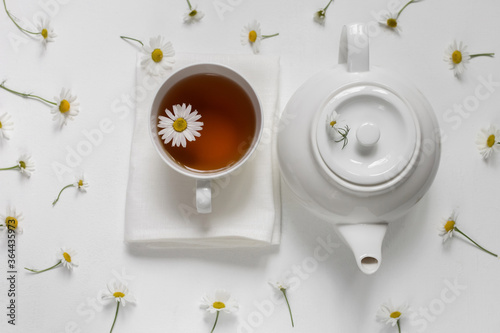 White cup with healthy natural chamomile tea on a white background near a white teapot and chamomile